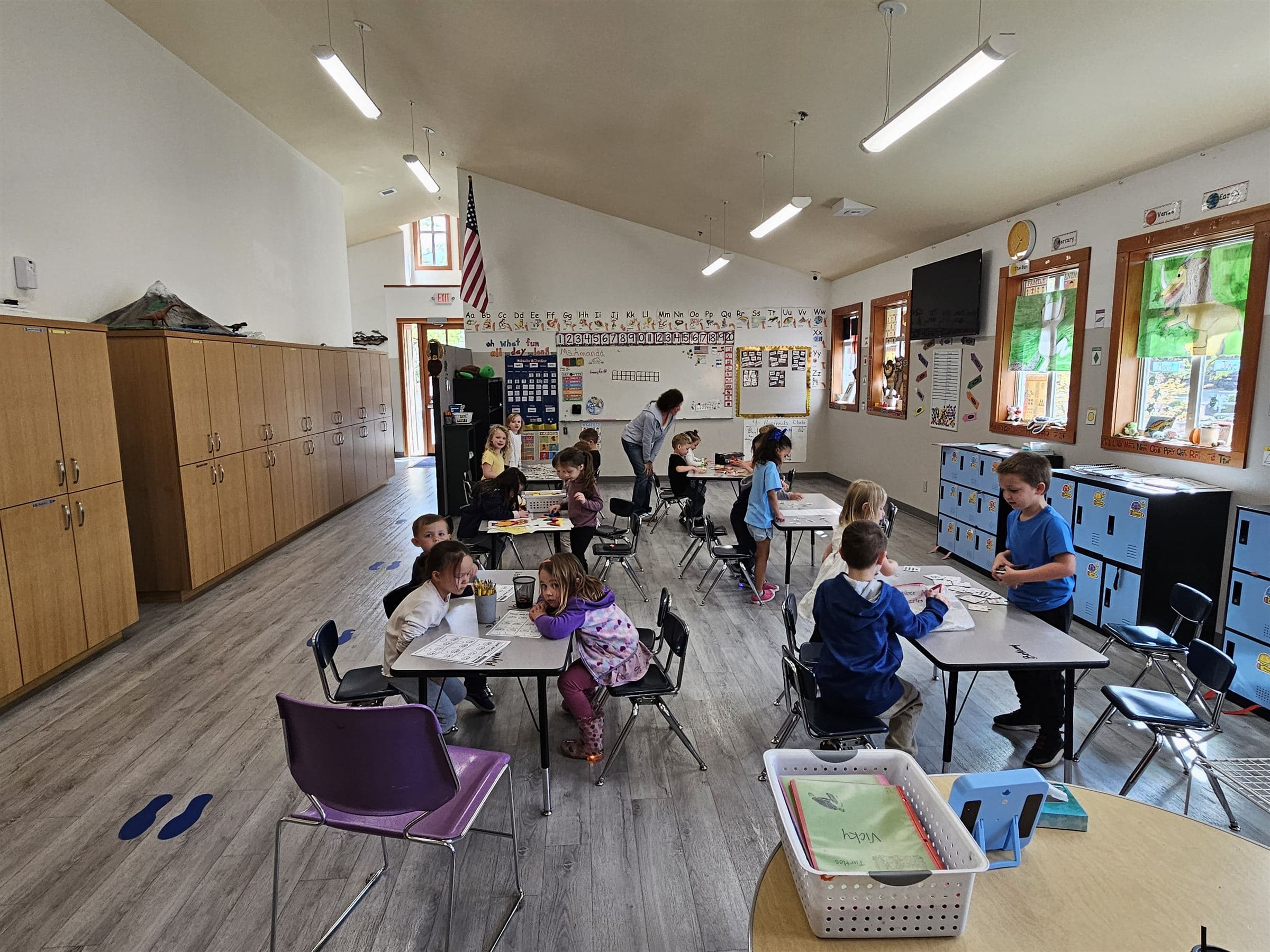 A bright 3rd–5th grade classroom with students working at tables while a teacher helps