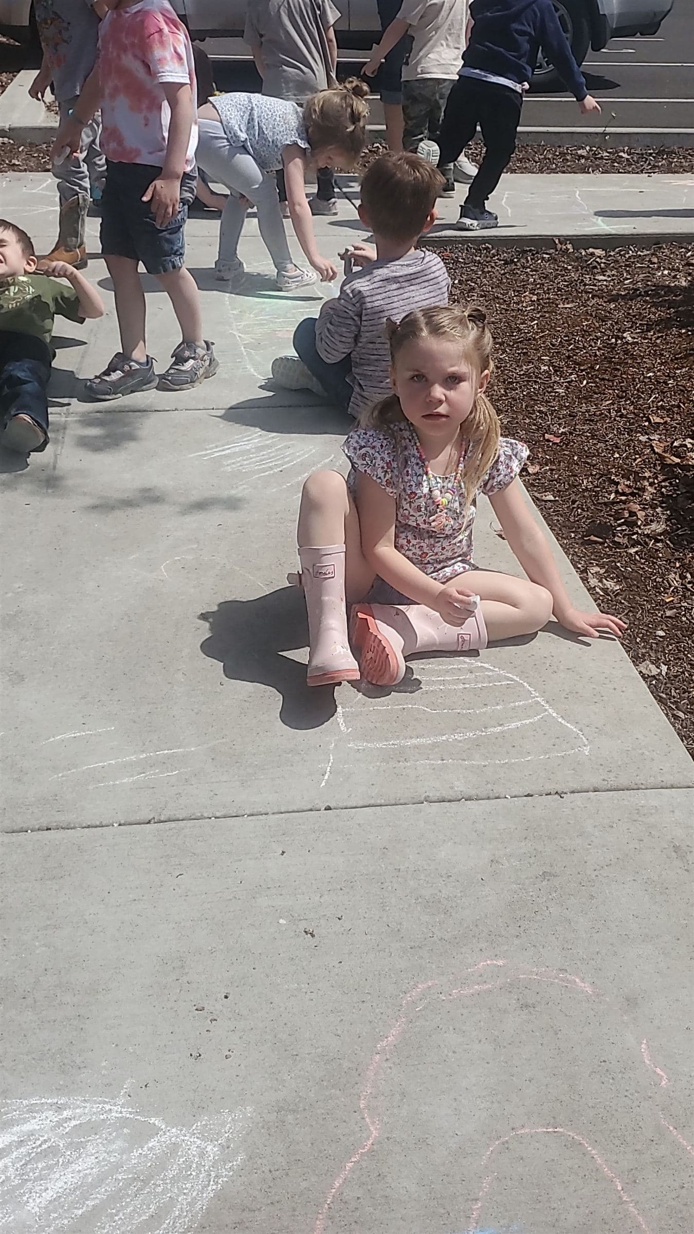 A kindergarten-age student sitting on the sidewalk surrounded by colorful chalk drawings, smiling in the sun