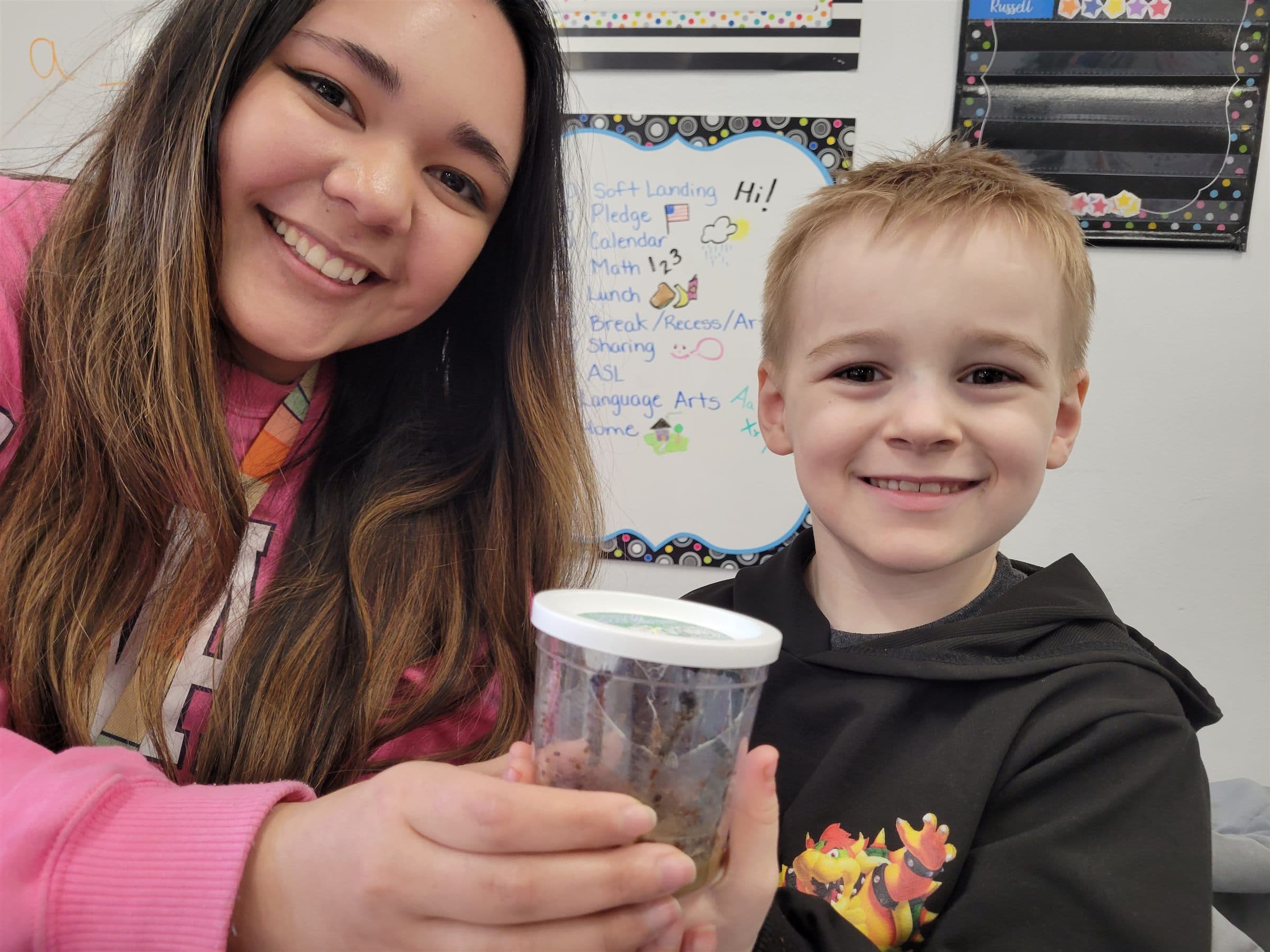 A teacher and a young student smiling together, holding up the caterpillar cup they're raising
